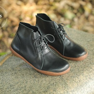 May include: A pair of black leather ankle boots with brown soles and laces. The boots are on a grey stone surface.