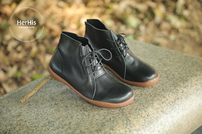 May include: A pair of black leather ankle boots with brown soles and laces. The boots are on a grey stone surface.