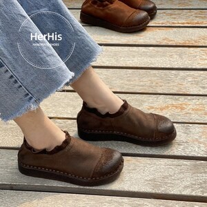 Op de afbeelding: Bruine leren instappers met een ronde neus en een gerimpelde kraag. De schoenen zijn handgemaakt en hebben een donkerbruine zool. De afbeelding bevat de tekst "HerHis HANDMADE SHOES".