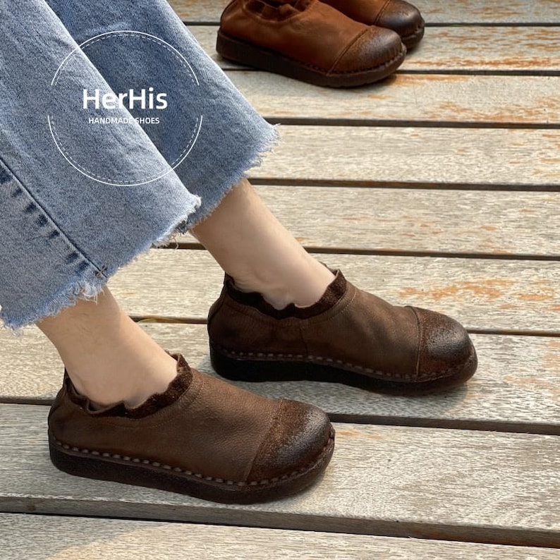 Op de afbeelding: Bruine leren instappers met een ronde neus en een gerimpelde kraag. De schoenen zijn handgemaakt en hebben een donkerbruine zool. De afbeelding bevat de tekst "HerHis HANDMADE SHOES".