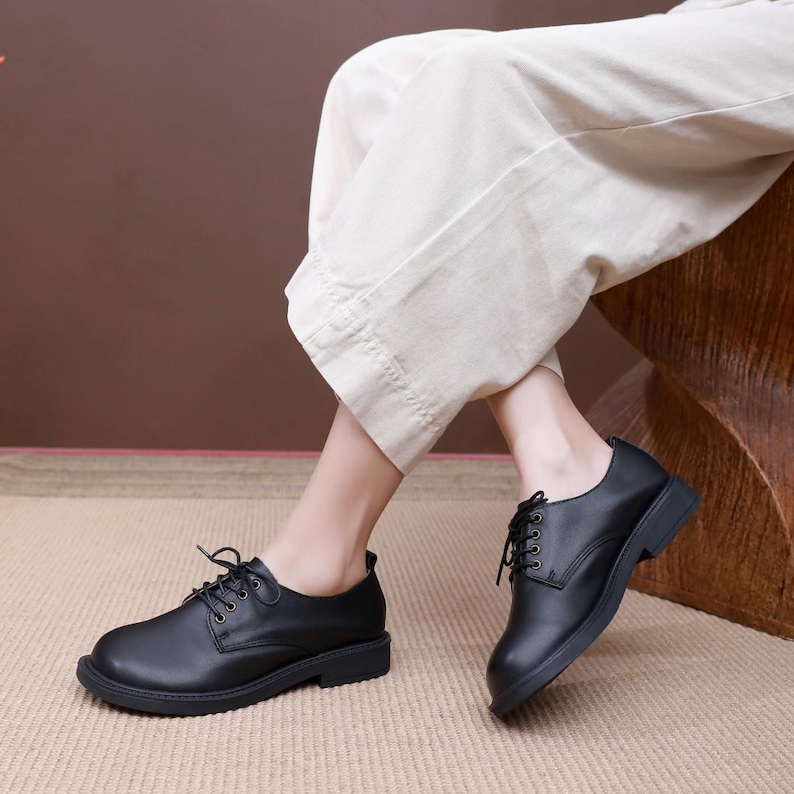 Puede incluir: Zapatos negros de cuero con cordones y suela gruesa. Los zapatos tienen un estilo oxford cl&aacute;sico y los lleva una persona con pantalones blancos.