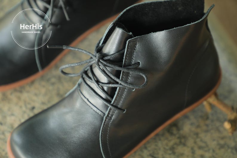 May include: Black leather ankle boots with laces. The boots have a rounded toe and a low heel. The brand name "HerHis" is visible on the side of the boot.