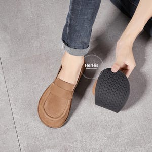 May include: A pair of brown leather loafers with a suede finish. The loafers have a simple design with a rounded toe and a flat sole. The soles are made of a textured rubber material. The loafers are shown on a person's feet, with the person's jeans visible in the background. The text "HerHis Handmade Shoes" is visible in the image.