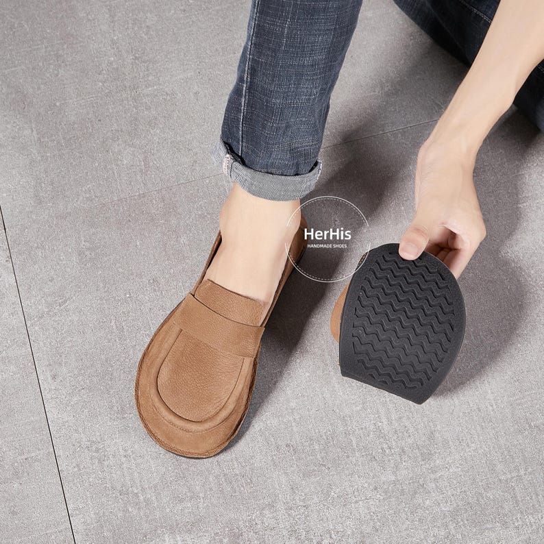 May include: A pair of brown leather loafers with a suede finish. The loafers have a simple design with a rounded toe and a flat sole. The soles are made of a textured rubber material. The loafers are shown on a person's feet, with the person's jeans visible in the background. The text "HerHis Handmade Shoes" is visible in the image.