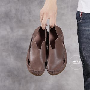 May include: A pair of brown leather slip-on shoes with a suede sole. The shoes have a simple design with a single strap across the top.