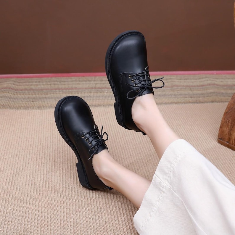 Puede incluir: Zapatos negros de cuero con cordones y suela gruesa. Los zapatos son usados por una persona con pantalones blancos.