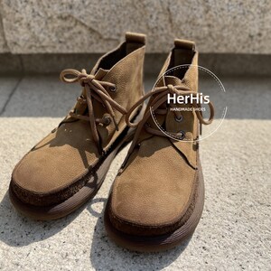May include: A pair of brown suede ankle boots with laces. The boots have a brown leather sole and a brown leather heel. The boots are handmade and have the text "HerHis HANDMADE SHOES" on the side.