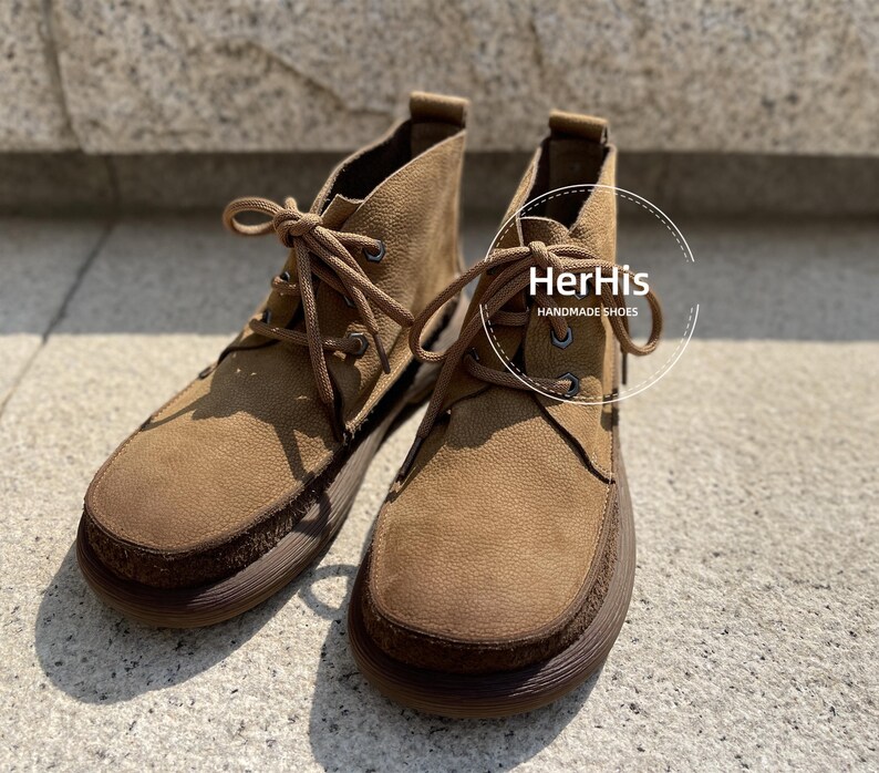 May include: A pair of brown suede ankle boots with laces. The boots have a brown leather sole and a brown leather heel. The boots are handmade and have the text "HerHis HANDMADE SHOES" on the side.