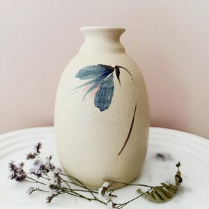 May include: A beige ceramic vase with a painted blue and pink floral design. The vase has a rounded body and a narrow neck. Dried purple flowers and a leaf are in front of the vase.