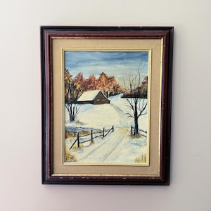 May include: An oil painting of a winter landscape featuring a snow-covered road leading to a cabin. The painting has a dark brown frame with a gold inner border. The sky is blue, and the trees have autumn colors.