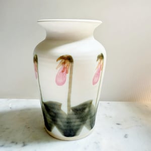 May include: A white ceramic vase with a wide base and a flared rim. The vase is decorated with pink and brown lady slipper orchid flowers and dark green leaves. The vase is approximately 25 cm tall.