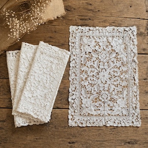 May include: Cream-colored lace placemat and three matching napkins. The rectangular placemat features a floral design and scalloped edges. The napkins are folded and have a similar lace pattern. The items are displayed on a rustic wooden surface.