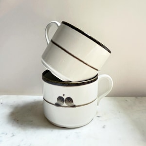 Peut inclure: Deux tasses en céramique blanches empilées. Chaque tasse a un bord marron foncé et une ligne diagonale marron. La tasse du bas présente un motif de deux oiseaux sur un fil.