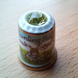 May include: A white porcelain thimble with a green gemstone on top. The thimble has a gold trim and a colorful illustration of a child hunting Easter eggs. The text "Hunting the Easter Eggs" is printed on the thimble.