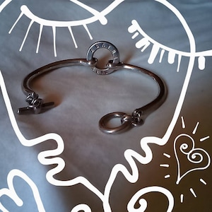Peut inclure: Un bracelet en argent avec un charme circulaire qui dit "Agatha Paris". Le charme a un petit cercle au centre. Le bracelet est ouvert et a une fermeture à crochet.
