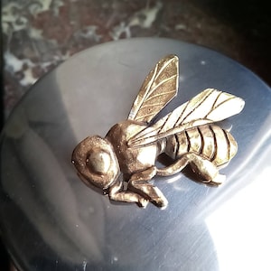 May include: A detailed, gold-colored bee figurine with textured wings and body, resting on a silver-colored, rounded surface. The bee's wings are outstretched, and the body has segmented details.