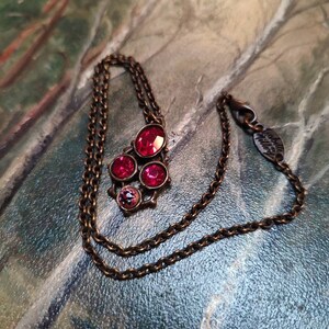 Peut inclure: Un collier en chaîne bronze avec un pendentif composé de quatre pierres de cristal rouges. Le pendentif a la forme d'un bouquet de fleurs. Le collier porte une petite étiquette avec le texte "Swarovski" dessus.