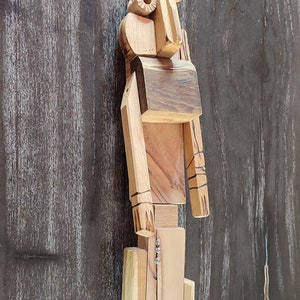 Puede incluir: Una escultura de madera de un robot con un diseño simple. El robot está hecho de diferentes piezas de madera y tiene una cabeza, brazos, piernas y un torso. El robot está pintado en un color madera natural.