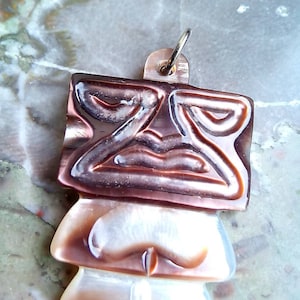Può includere: Un ciondolo in conchiglia marrone e bianco con un design intagliato a forma di viso tiki. Il ciondolo ha un anello d'argento per appenderlo.