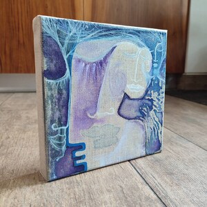 Può includere: Dipinto astratto su tela con una gamma di colori viola e blu. Il dipinto presenta due volti, uno con gli occhi chiusi e l'altro con gli occhi aperti. I volti sono parzialmente oscurati da un motivo blu e bianco.