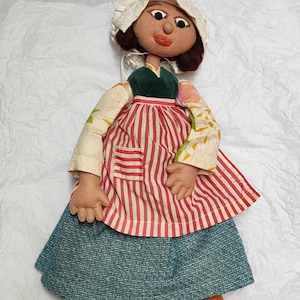 Könnte beinhalten: Eine Vintage-Handpuppe mit einem weißen Häubchen, einem grünen Oberteil, einer rot-weiß gestreiften Schürze und einem blauen Rock. Die Puppe trägt Holzschuhe mit orange-blau-gelb gestreiften Socken.