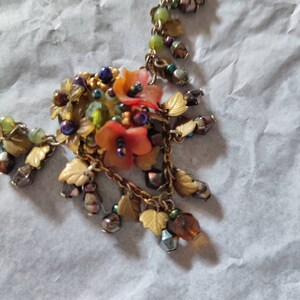 Può includere: Una collana d'oro con un ciondolo floreale con perline arancioni, verdi e viola. Il ciondolo è circondato da foglie e perline dorate.