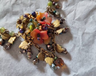 May include: A gold necklace with a floral pendant featuring orange, green, and purple beads. The pendant is surrounded by gold leaves and beads.