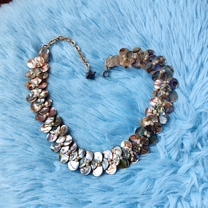 Pode incluir: Um colar feito de peças de concha iridescente multicoloridas, cada uma com um pequeno detalhe de pérola. O colar é conectado por uma corrente de ouro.