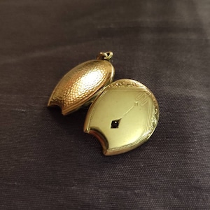 Può includere: Un medaglione d'oro con una superficie testurizzata e un coperchio incernierato. Il medaglione è aperto, rivelando un interno liscio in oro con un piccolo design a forma di diamante nero.