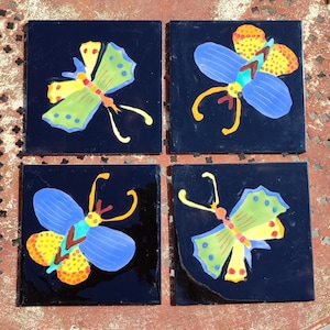 Puede incluir: Cuatro baldosas de cerámica cuadradas con acabado negro brillante. Cada baldosa presenta un diseño de mariposa colorido con detalles en azul, amarillo, verde y naranja. Las alas de la mariposa tienen un patrón punteado.