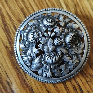 Op de afbeelding: Een gedetailleerde, zilverkleurige bloemenbroche. De ronde broche heeft een centrale opstelling van gebeeldhouwde bloemen en bladeren, omgeven door een kralenrand. Het ontwerp toont ingewikkelde details en een vintage esthetiek, geschikt om kleding te versieren.
