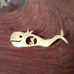 May include: A gold-colored whale brooch with a cutout design featuring a person. The brooch has a pin on the back for easy attachment. The whale's body is smooth, and the design is whimsical.