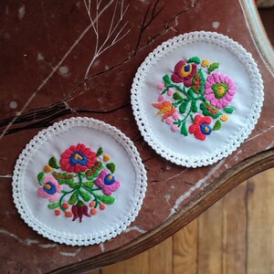 Könnte beinhalten: Zwei weiße Häkeluntersetzer mit gestickten Blumenmustern. Die Blumen sind rosa, rot, grün und gelb. Die Untersetzer befinden sich auf einer braunen Marmorfläche.