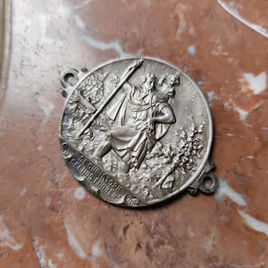 Könnte beinhalten: Silberfarbene runde Medaille mit einem detaillierten Relief von St. Christophorus, der ein Kind durch das Wasser trägt. Der Text "ST. CHRISTOPH SEI UNS FUHRER!" ist am Rand eingeschrieben. Die Medaille hat zwei kleine Schlaufen zum Aufhängen.
