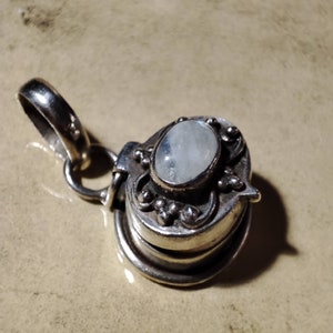 Può includere: Ciondolo in argento con una pietra ovale di colore chiaro incastonata in un design ornato e stratificato. Il ciondolo ha un vano incernierato e un anello per una catena. Il metallo ha una finitura lucida e antica.