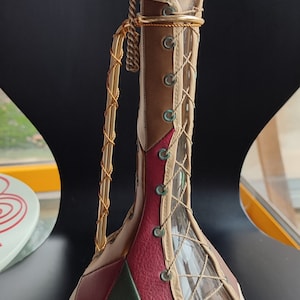 May include: A glass bottle with a leather covering. The bottle is decorated with a patchwork of brown, red, and green leather. The bottle has a cork stopper and a gold chain handle.