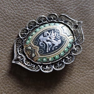 May include: An ornate silver brooch with an oval, black enamel center featuring a silver mythical creature. The brooch has a filigree border and is accented with green and gold details. The overall design is intricate and detailed.