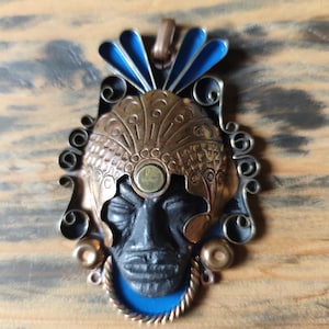May include: A detailed pendant featuring a black face with closed eyes, adorned with a copper-toned headdress and blue accents. The pendant has a silver-colored frame and a blue base, creating a striking contrast. The pendant is likely made of metal.
