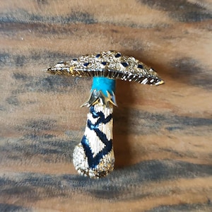 Pode incluir: Broche dourado em forma de cogumelo com uma tampa texturizada adornada com detalhes em azul escuro. O caule apresenta um padrão em zigue-zague preto e dourado, e as brânquias são de um turquesa vibrante. O broche está sobre um fundo castanho.