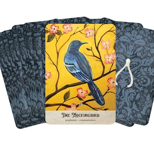 Puede incluir: Un mazo de cartas de tarot con un p&aacute;jaro azul posado en una rama con flores rosas sobre un fondo amarillo. La carta dice "The Mockingbird" con las palabras "prophecies" y "communication" debajo. Los dorsos de las cartas tienen un patr&oacute;n floral azul oscuro.