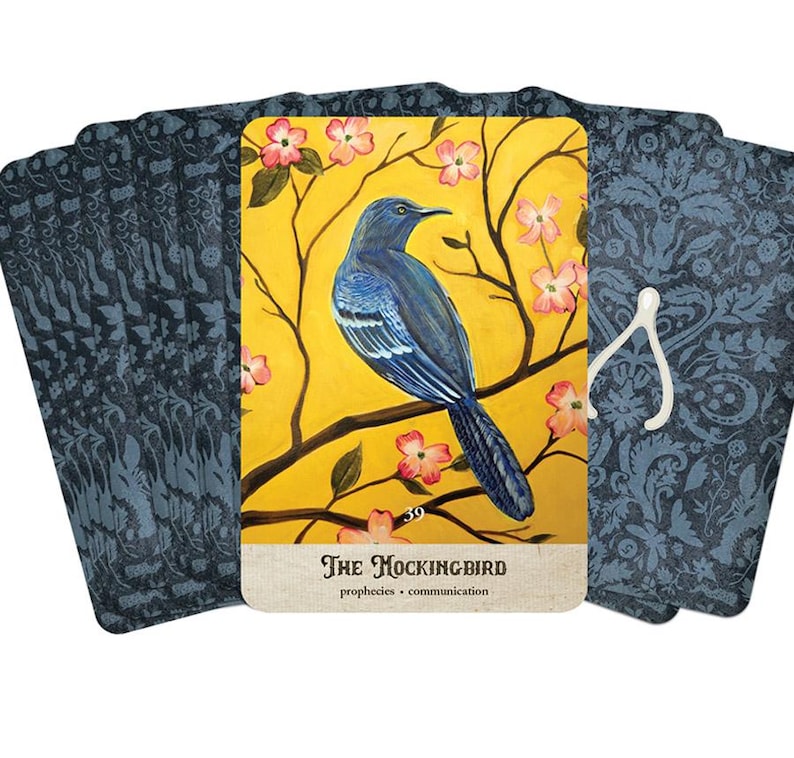 Puede incluir: Un mazo de cartas de tarot con un p&aacute;jaro azul posado en una rama con flores rosas sobre un fondo amarillo. La carta dice "The Mockingbird" con las palabras "prophecies" y "communication" debajo. Los dorsos de las cartas tienen un patr&oacute;n floral azul oscuro.