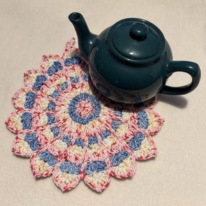 May include: A teal ceramic teapot rests on a handmade, colorful crochet coaster. The coaster features a floral design with red, white, blue, and yellow yarn. The teapot has a rounded body, a curved handle, and a spout.