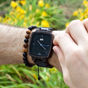 K&ouml;nnte beinhalten: Eine schwarze Holz-Armbanduhr mit schwarzem Zifferblatt und silbernen Zeigern. Die Uhr hat ein schwarzes Lederband und ein braunes Holzperlenarmband.