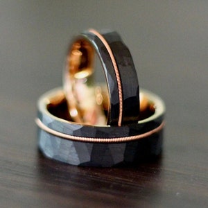 May include: Two black and rose gold wedding bands with a hammered finish. The bands are stacked on top of each other.