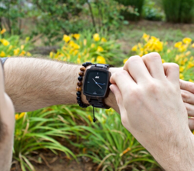 K&ouml;nnte beinhalten: Eine schwarze Holz-Armbanduhr mit schwarzem Zifferblatt und silbernen Zeigern. Die Uhr hat ein schwarzes Lederband und ein braunes Holzperlenarmband.