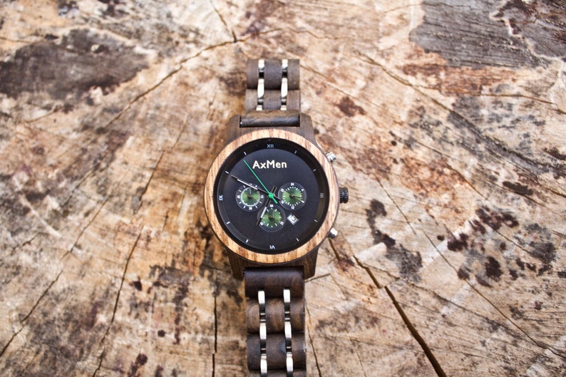 Puede incluir: Un reloj de pulsera de madera negro y marr&oacute;n con una esfera de cron&oacute;grafo. La esfera del reloj tiene un fondo negro con marcas blancas y acentos verdes. El nombre de la marca "AxMen" est&aacute; impreso en la esfera del reloj.