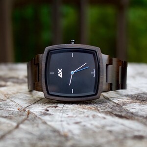 K&ouml;nnte beinhalten: Eine quadratische Holzuhr mit schwarzem Zifferblatt und silbernen Zeigern. Die Uhr hat ein dunkelbraunes Holzband.
