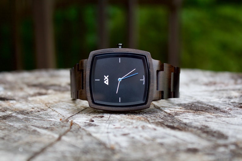 K&ouml;nnte beinhalten: Eine quadratische Holzuhr mit schwarzem Zifferblatt und silbernen Zeigern. Die Uhr hat ein dunkelbraunes Holzband.