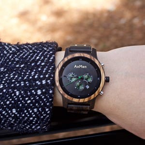 Puede incluir: Un reloj de pulsera de madera negro y marr&oacute;n con una esfera de cron&oacute;grafo. La esfera del reloj tiene agujas blancas y detalles verdes. La esfera del reloj tambi&eacute;n tiene el texto "AxMen" impreso en ella.