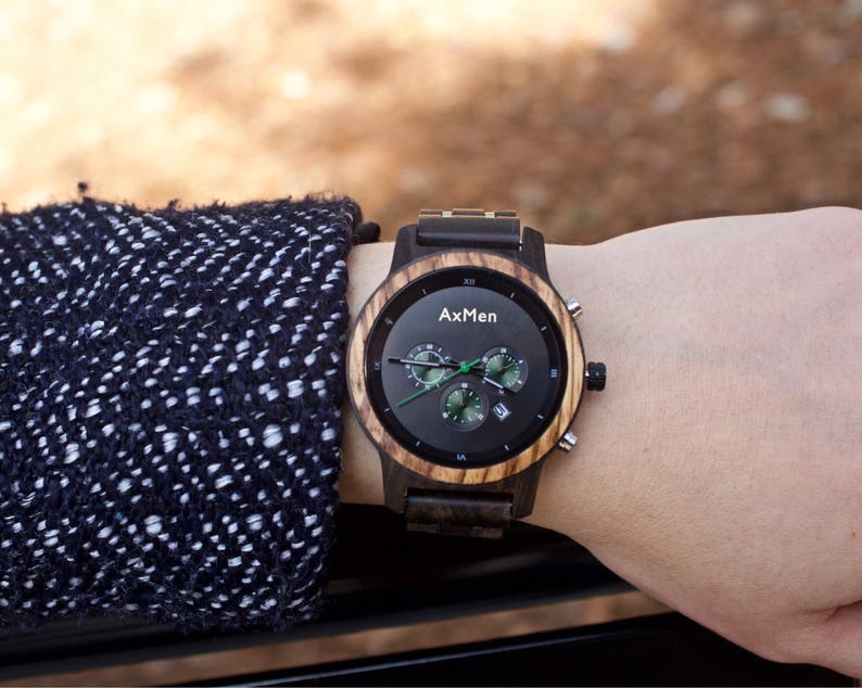 Puede incluir: Un reloj de pulsera de madera negro y marr&oacute;n con una esfera de cron&oacute;grafo. La esfera del reloj tiene agujas blancas y detalles verdes. La esfera del reloj tambi&eacute;n tiene el texto "AxMen" impreso en ella.